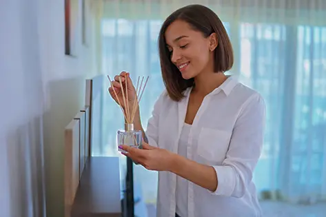 Woman placing reed sticks into a glass diffuser in a calm, sunlit living room