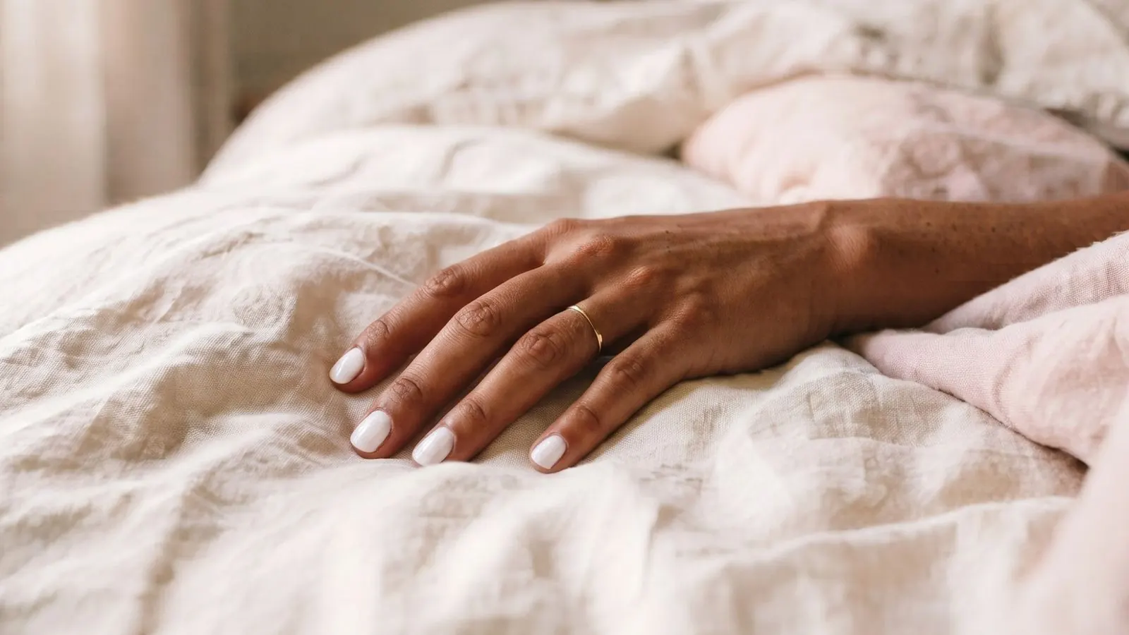 A woman's hand with a thin gold ring resting on soft cream and blush linen bedding in warm morning light